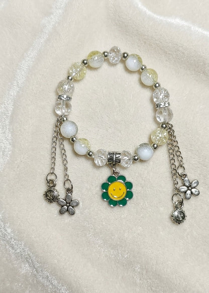 A beaded charm bracelet with white and pale yellow beads, silver-toned metal chains, and a central green and yellow flower charm, displayed on a soft cream-colored fabric background.