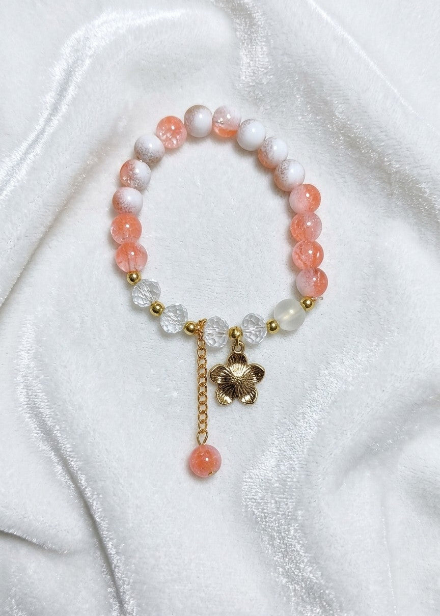 A beaded stretch bracelet featuring translucent peach-orange and white beads, accented with gold-tone spacer beads, clear faceted crystal flower charms, a gold-tone flower charm, and a dangling peach-orange bead, displayed on a white satin fabric background.