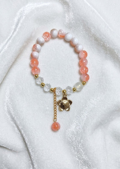 A beaded stretch bracelet featuring translucent peach-orange and white beads, accented with gold-tone spacer beads, clear faceted crystal flower charms, a gold-tone flower charm, and a dangling peach-orange bead, displayed on a white satin fabric background.