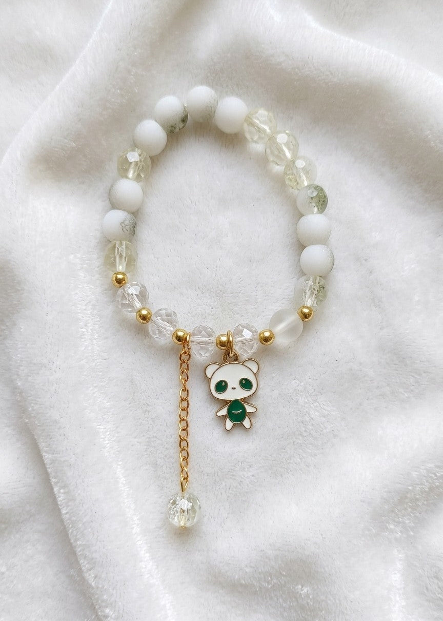 A beaded stretch bracelet featuring white and clear faceted beads, small gold spacer beads, and a dangling charm with a white panda face and green chest detail, displayed on a soft white fabric background.