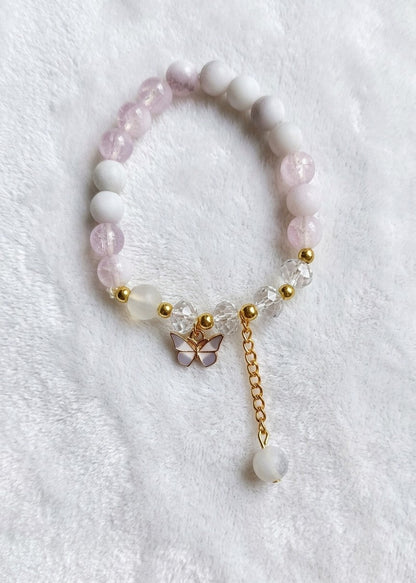 A beaded bracelet with alternating pale pink and white round beads, accented by small gold spacer beads, a butterfly charm, and a dangling gold chain with a white bead, laid on a white textured fabric background.