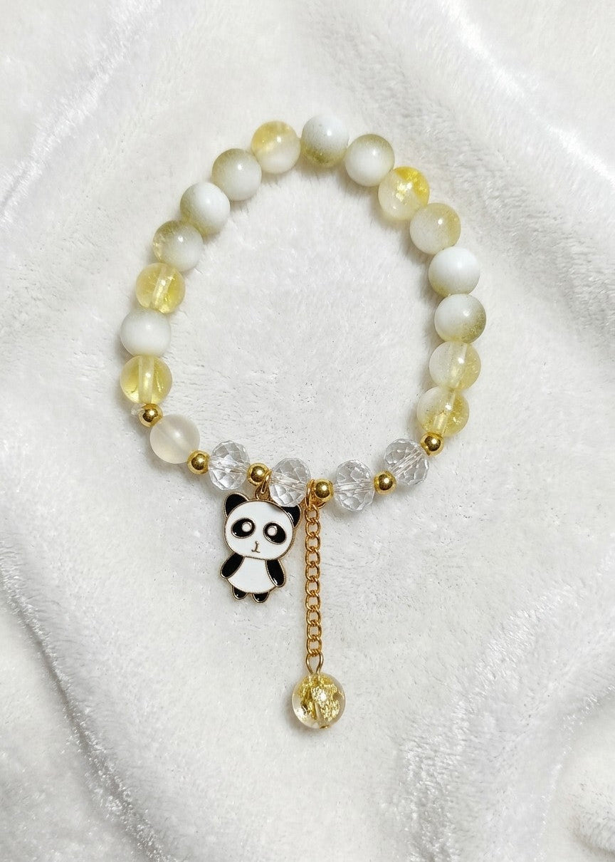 A beaded stretch bracelet featuring white and pale yellow translucent beads, gold spacer beads, a small black-and-white panda charm, and a dangling gold bead, displayed on a soft white fabric background.