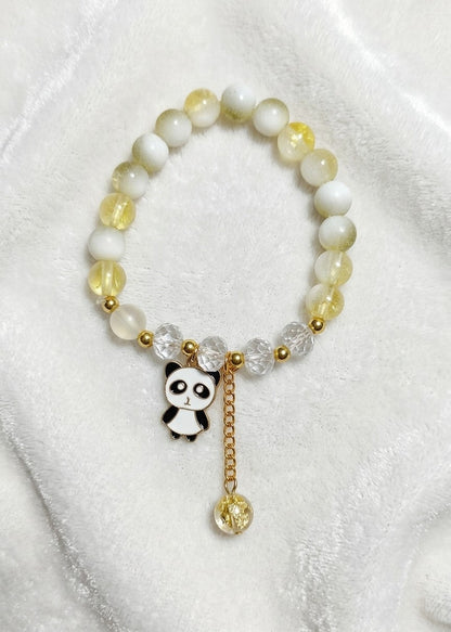 A beaded stretch bracelet featuring white and pale yellow translucent beads, gold spacer beads, a small black-and-white panda charm, and a dangling gold bead, displayed on a soft white fabric background.
