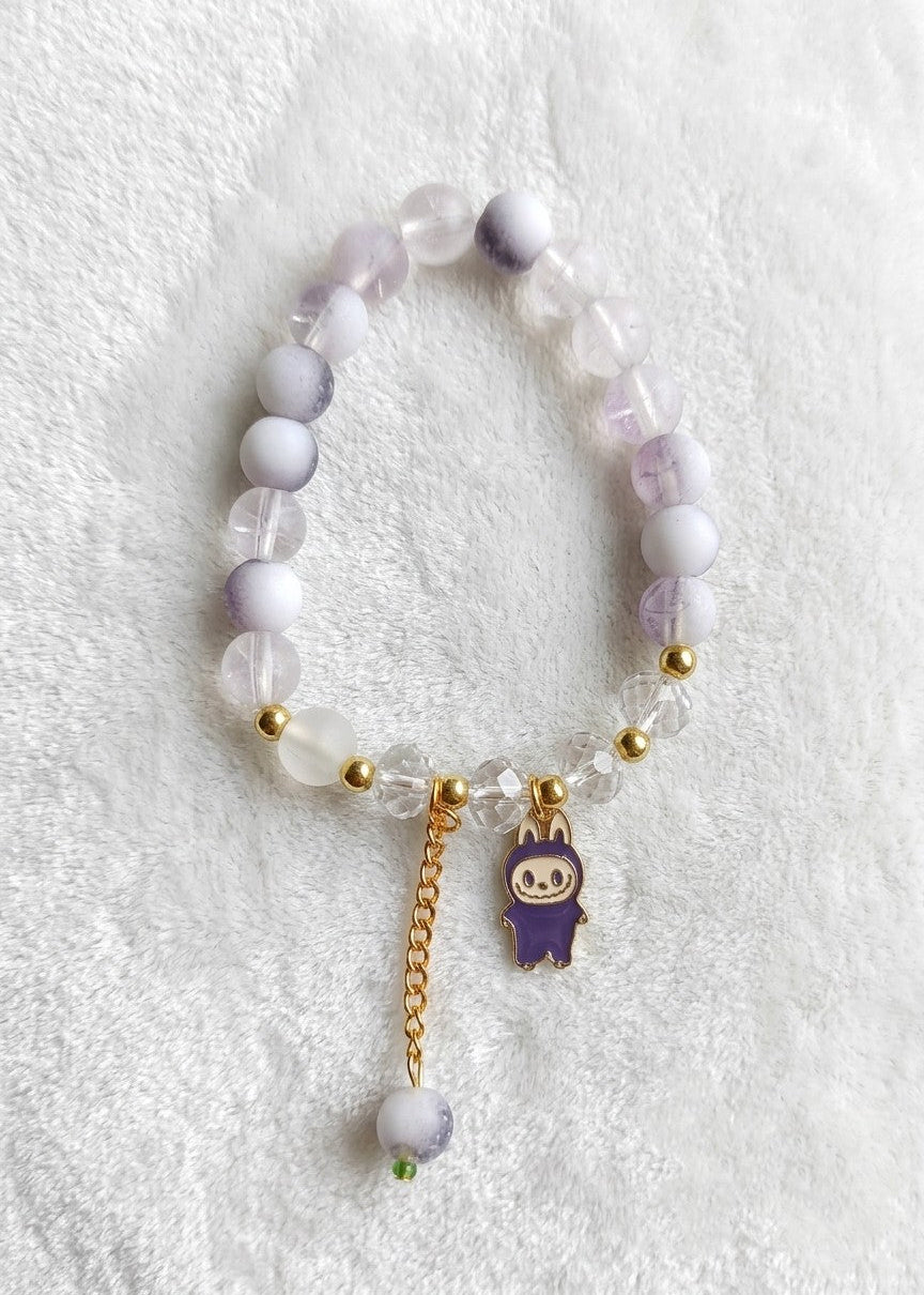 A beaded stretch bracelet featuring pale lavender and white translucent beads, small gold spacer beads, a gold chain, and a purple labubu character charm, displayed on a white textured fabric background.