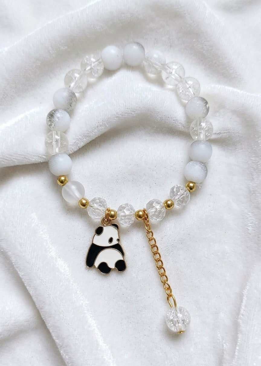 A beaded bracelet featuring white opaque beads, clear faceted beads, small gold spacer beads, and a dangling charm shaped like a black-and-white panda with a gold chain and clear bead drop, displayed on a white fabric background.