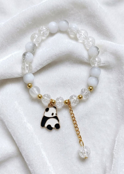 A beaded bracelet featuring white opaque beads, clear faceted beads, small gold spacer beads, and a dangling charm shaped like a black-and-white panda with a gold chain and clear bead drop, displayed on a white fabric background.