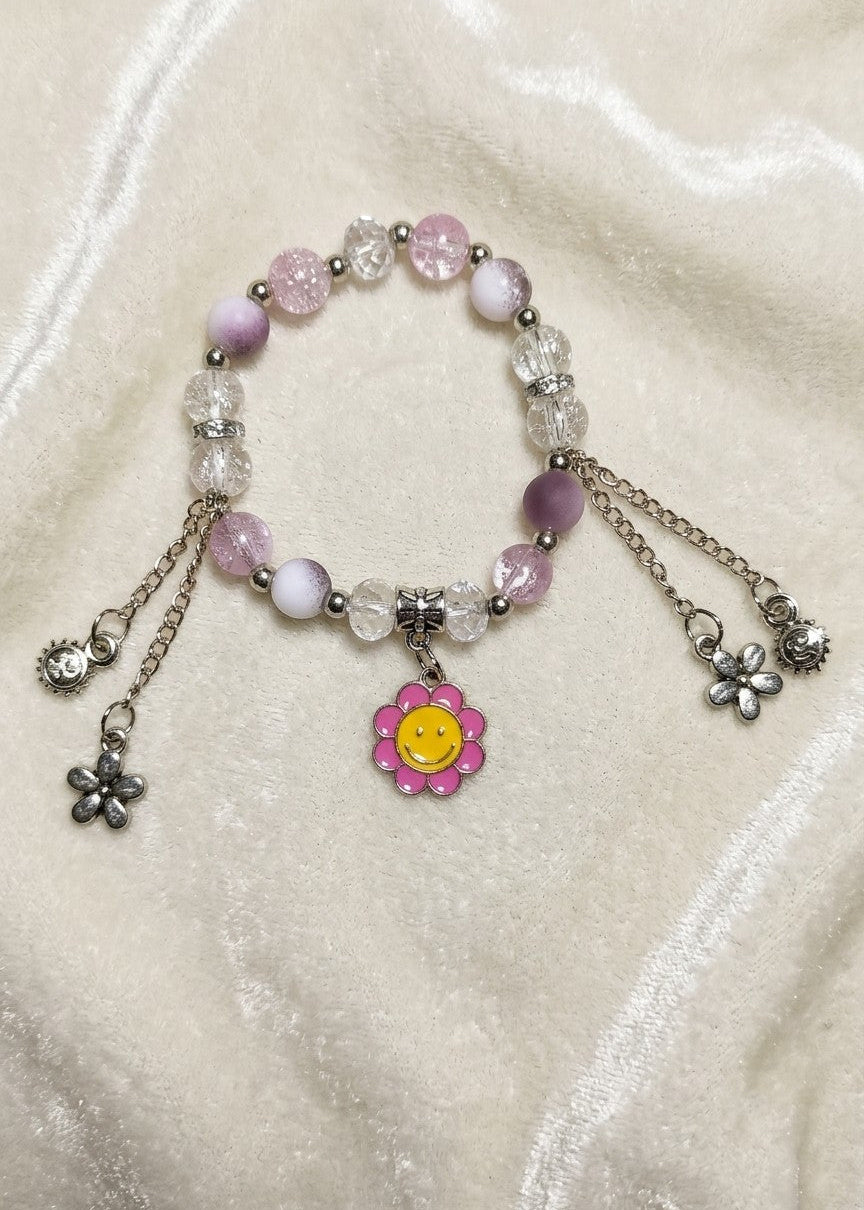 A beaded charm bracelet with pink and white beads, clear faceted beads, silver-toned chains, and dangling flower charms, including a central pink flower charm with a yellow smiley face, displayed on a cream satin fabric background.