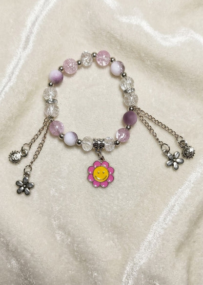 A beaded charm bracelet with pink and white beads, clear faceted beads, silver-toned chains, and dangling flower charms, including a central pink flower charm with a yellow smiley face, displayed on a cream satin fabric background.