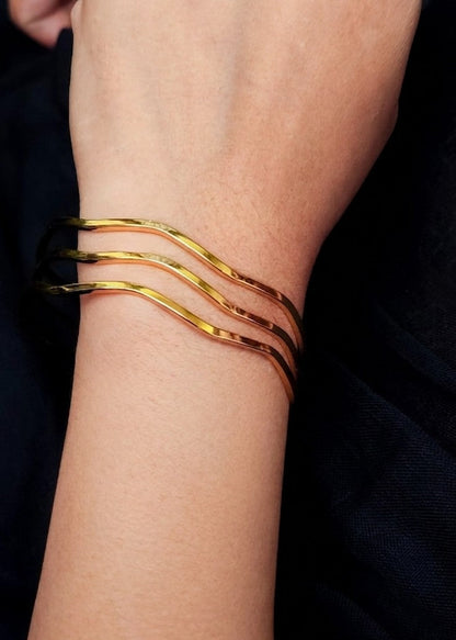 Three thin wavy gold bangles stacked together on a person's wrist, shown against a dark navy or black fabric background.