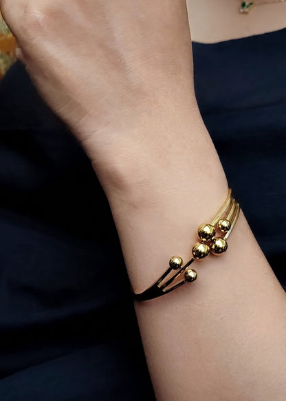 A close-up of a person's wrist wearing a gold-toned multi-strand bracelet with small polished bead details and a black wrap band.
