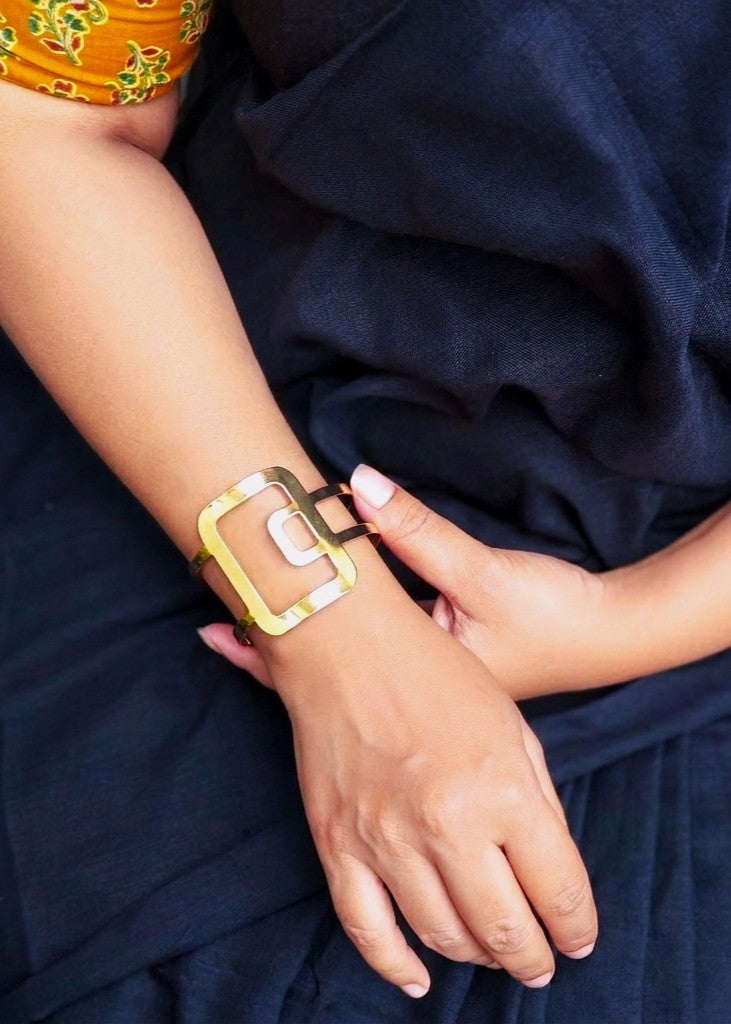 A close-up of a person's wrist wearing a chunky, geometric gold-toned cuff bracelet with a large square frame and a central rectangular detail, set against a dark navy blue fabric background.