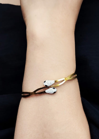 A close-up of a person's wrist wearing a gold and dark brown bangle bracelet with two snake head motifs set with clear crystals.