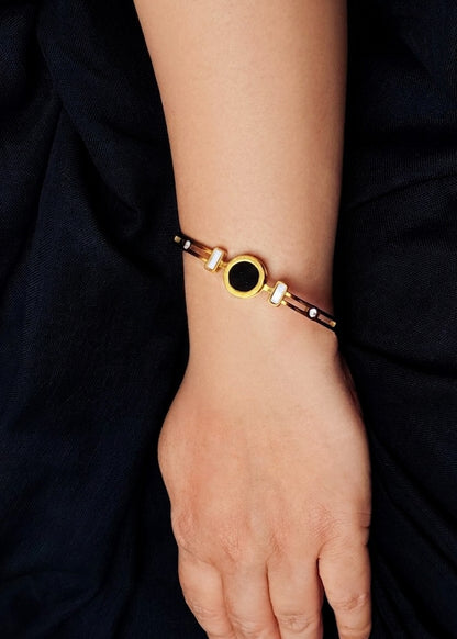 A close-up of a person's wrist wearing a gold-toned bangle bracelet with a central black oval stone and small white accent stones, set against dark navy fabric.