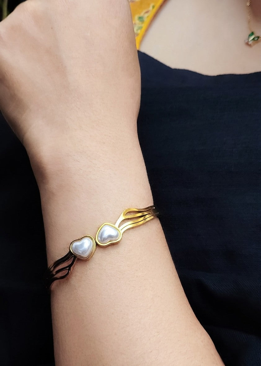 A close-up of a person's wrist wearing a gold-toned multi-strand bracelet with two heart-shaped pearl accents and a thin black cord detail.