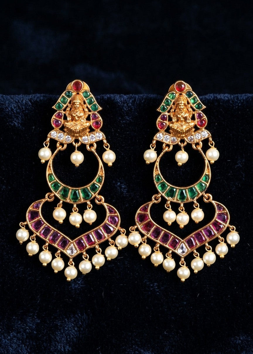 A pair of ornate gold-plated Indian chandbali earrings decorated with green and purple enamel, red stones, and white faux pearls, displayed on a dark velvet background.