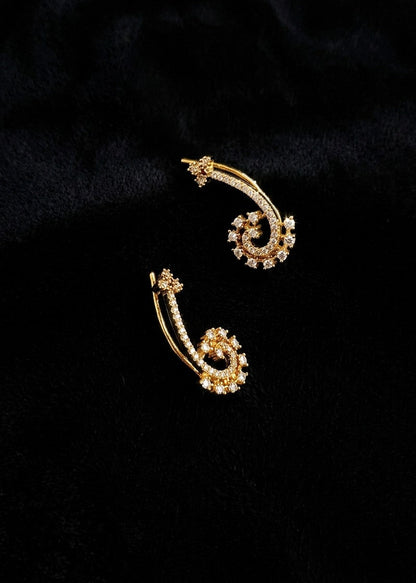 A pair of gold-toned hoop earrings with intricate spiral details and small clear stones, displayed on a textured black fabric background.