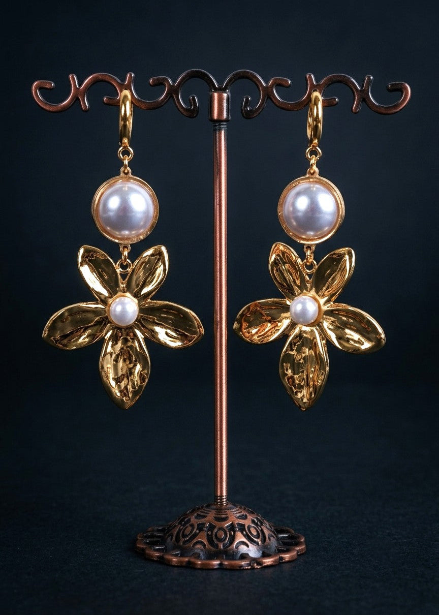 A pair of gold flower-shaped dangle earrings with round white pearl-like beads at the top and a small white pearl at the center of each flower, displayed on a copper jewelry stand against a dark background.
