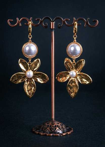 A pair of gold flower-shaped dangle earrings with round white pearl-like beads at the top and a small white pearl at the center of each flower, displayed on a copper jewelry stand against a dark background.