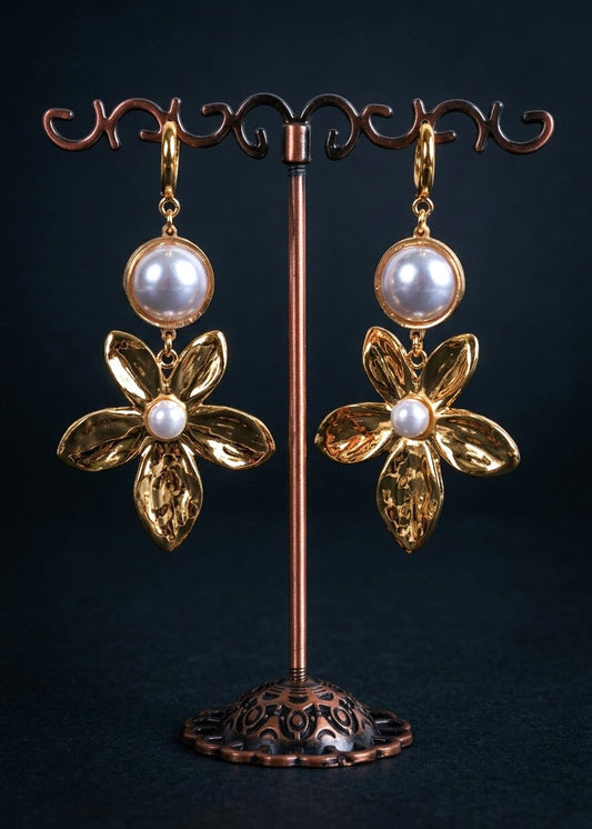 A pair of gold flower-shaped dangle earrings with round white pearl-like beads at the top and a small white pearl at the center of each flower, displayed on a copper jewelry stand against a dark background.