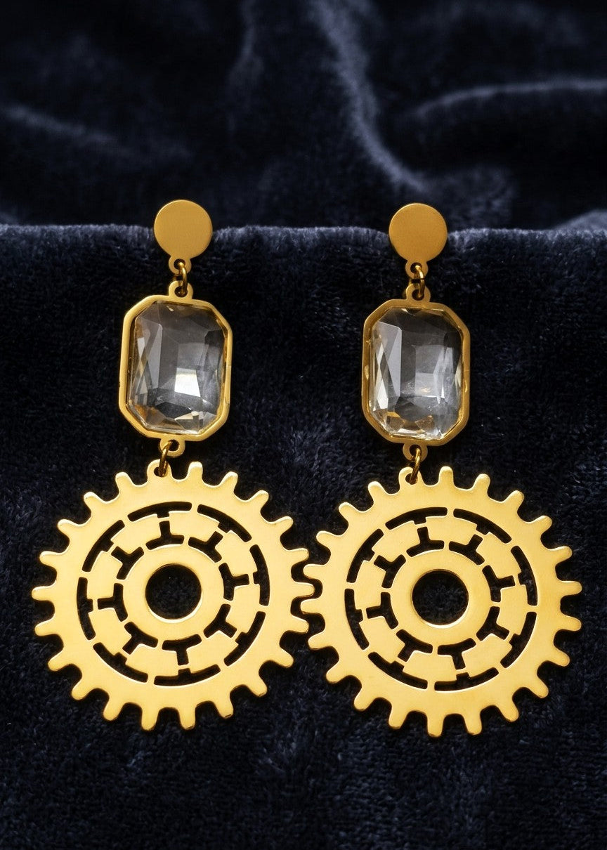 Steampunk-style drop earrings with gold-tone gear-shaped charms and clear rectangular gemstones, displayed on a dark velvet surface.