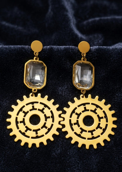 Steampunk-style drop earrings with gold-tone gear-shaped charms and clear rectangular gemstones, displayed on a dark velvet surface.