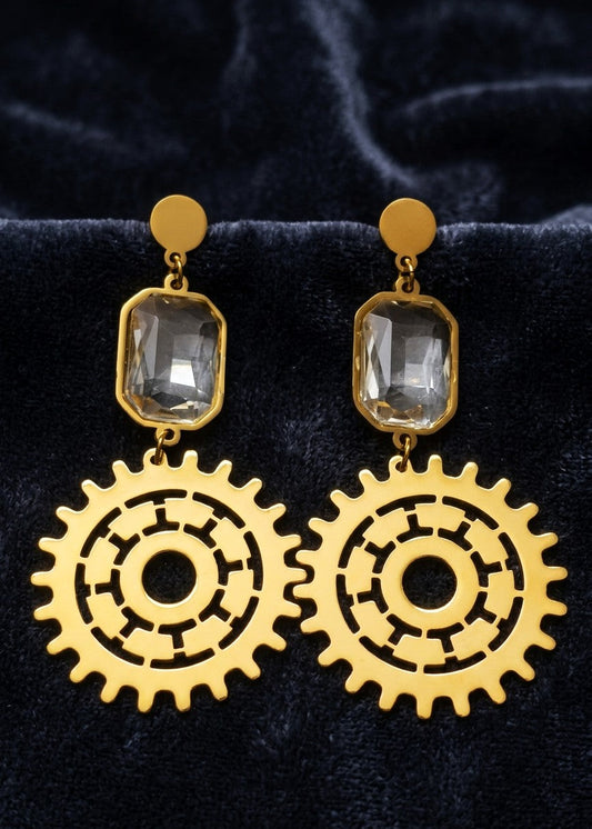 Steampunk-style drop earrings with gold-tone gear-shaped charms and clear rectangular gemstones, displayed on a dark velvet surface.