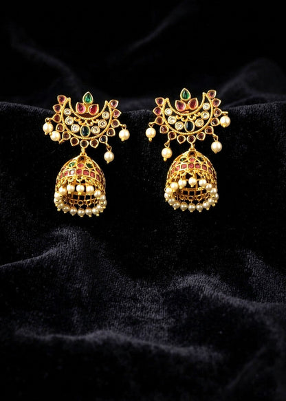 A pair of ornate gold-toned jhumka earrings with red and green enamel details, set against a black velvet background.