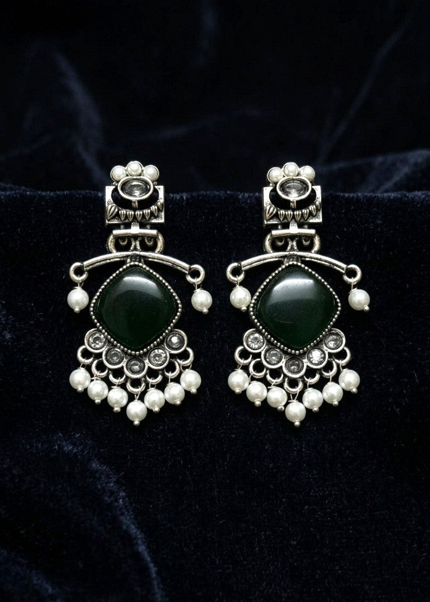 A pair of silver-toned traditional Indian jhumka earrings with dark green stone centers and small white pearl-like beads, displayed on a black velvet background.