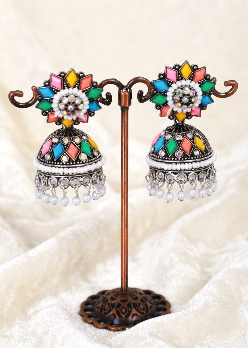 A pair of traditional Indian jhumka earrings displayed on a decorative bronze stand. The earrings feature colorful enamel details in pink, yellow, green, and blue, white beadwork, and silver-toned dangling charms, set against a soft beige fabric background.