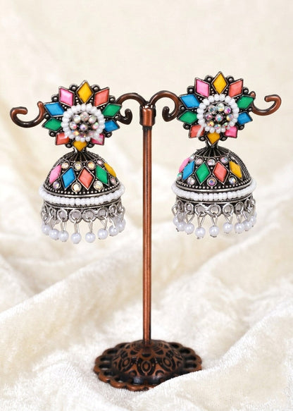 A pair of traditional Indian jhumka earrings displayed on a decorative bronze stand. The earrings feature colorful enamel details in pink, yellow, green, and blue, white beadwork, and silver-toned dangling charms, set against a soft beige fabric background.