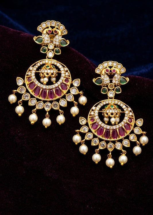 A pair of ornate gold-toned Indian jhumka earrings decorated with white pearls, red stones, and green stones, displayed on a dark velvet background.