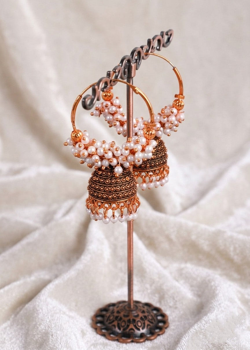 A pair of traditional Indian jhumka earrings displayed on a decorative stand. The earrings feature copper-toned hoops adorned with white pearl bead clusters and dangling copper-toned jhumka bells with intricate filigree work and pearl bead borders.