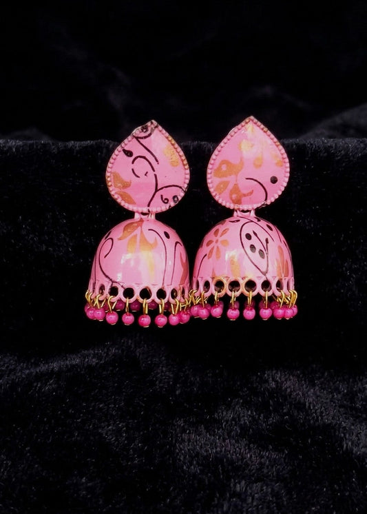 A pair of pink jhumka earrings with gold and orange painted patterns, displayed on a black velvet background.