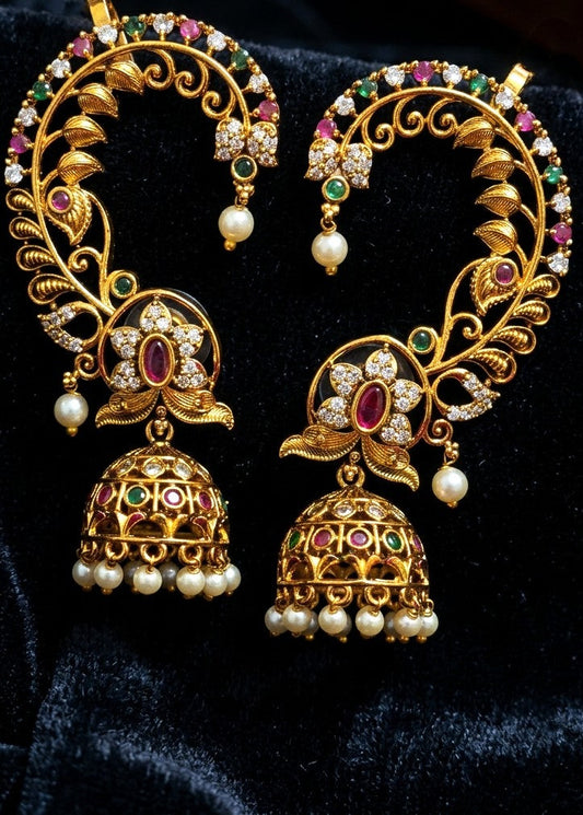 A pair of ornate gold-toned traditional Indian earrings with filigree, green and pink enamel stones, and dangling pearl beads, displayed on a black velvet background.