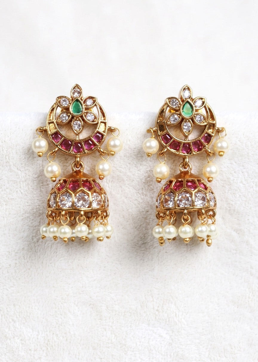 A pair of gold-plated Indian jhumka earrings decorated with green emeralds, pink rhinestones, and white faux pearls, displayed on a soft white textured fabric background.