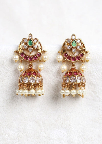 A pair of gold-plated Indian jhumka earrings decorated with green emeralds, pink rhinestones, and white faux pearls, displayed on a soft white textured fabric background.