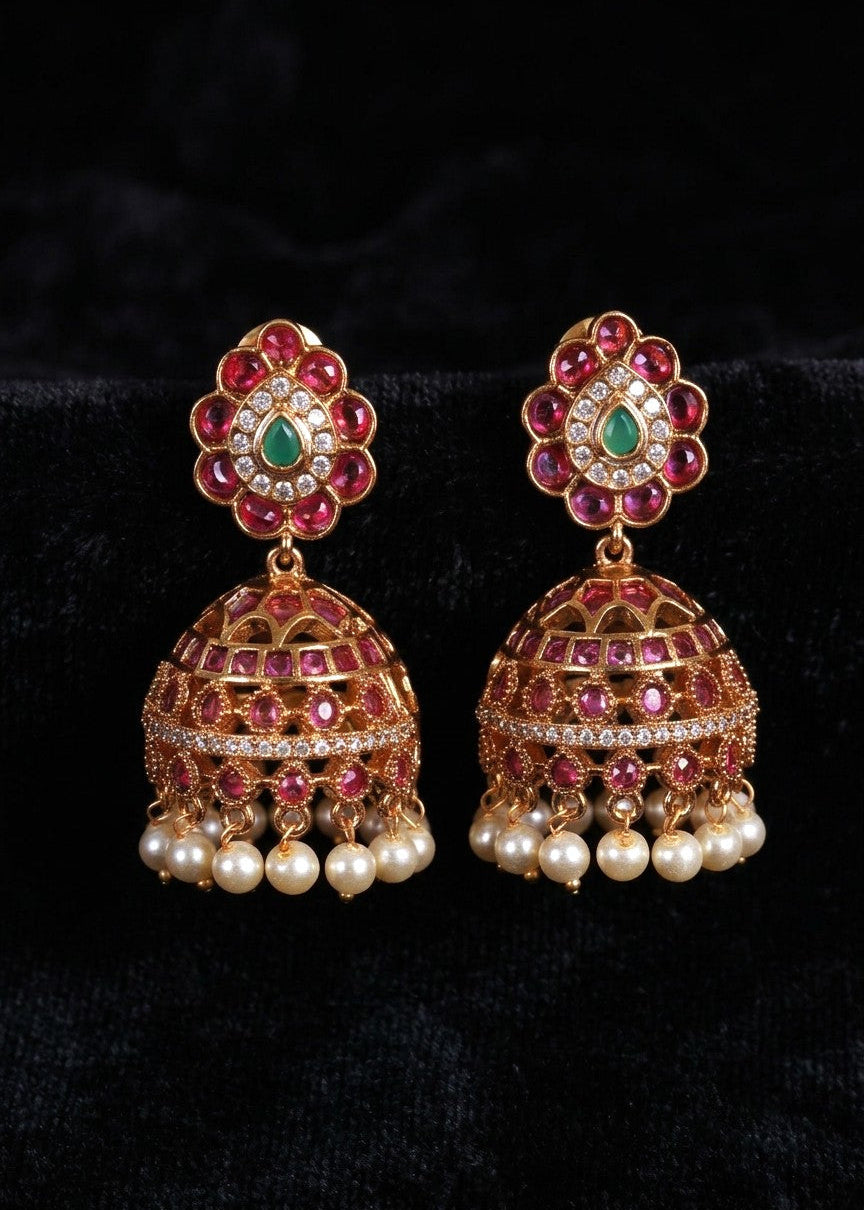 A pair of ornate gold-toned jhumka earrings with red gemstone accents, a central green stone, and dangling pearl beads, displayed on a black velvet background.
