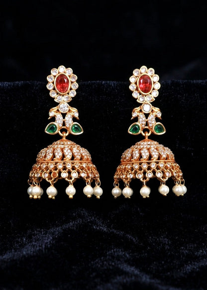 A pair of ornate gold-toned Indian jhumka earrings with red oval stones, green leaf accents, clear stones, and dangling pearl-like beads, displayed on a black velvet background.