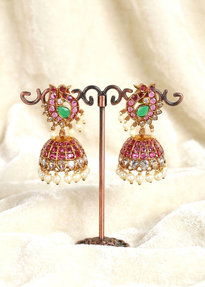 A pair of ornate gold-toned jhumka earrings with pink and green stones and white pearl bead drops, displayed on a decorative stand against a soft cream fabric background.