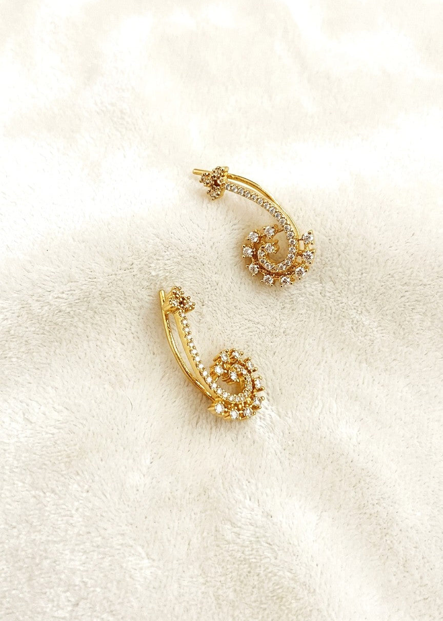 A pair of gold crescent-shaped hoop earrings adorned with small clear crystals, displayed on a soft, textured cream fabric background.