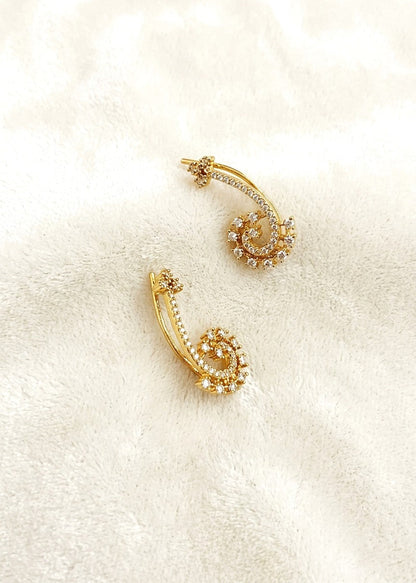 A pair of gold crescent-shaped hoop earrings adorned with small clear crystals, displayed on a soft, textured cream fabric background.