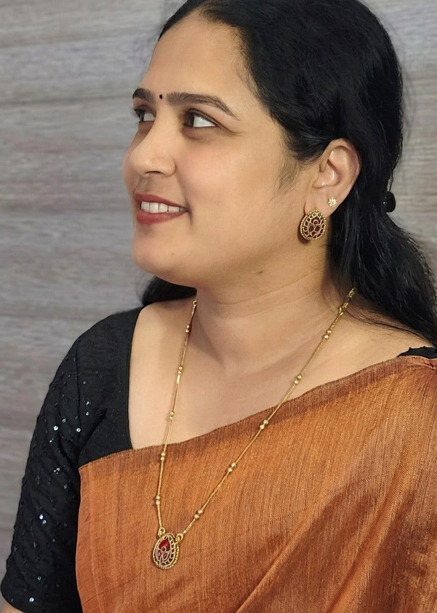 A woman in profile wearing a gold necklace with a red teardrop pendant, matching gold earrings, and a black blouse with a rust-orange sari, set against a gray textured background.