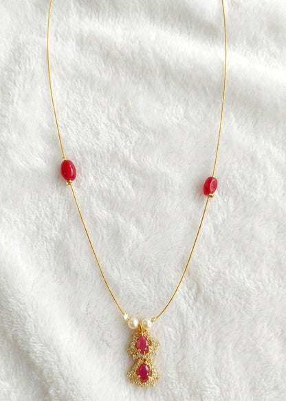 A delicate gold necklace featuring three small red oval beads on the chain and a central gold pendant with two red teardrop-shaped stones, displayed on a soft white textured fabric background.