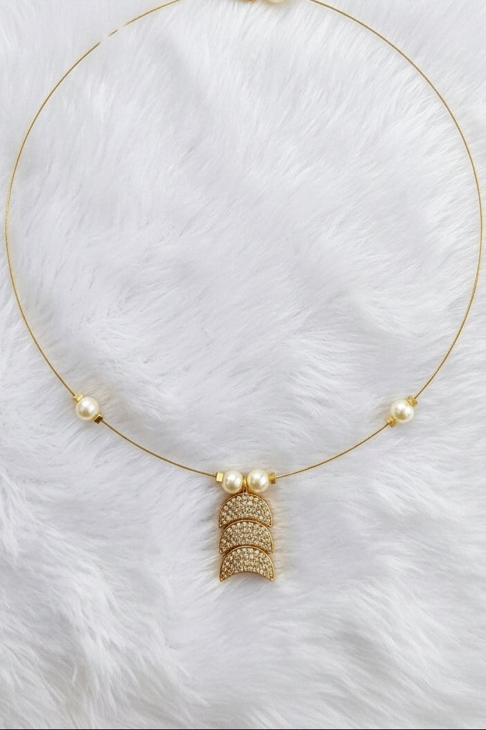 A delicate gold necklace with three small white pearls spaced along the chain and a central gold pendant featuring textured stacked crescent shapes, displayed on a white faux fur background.