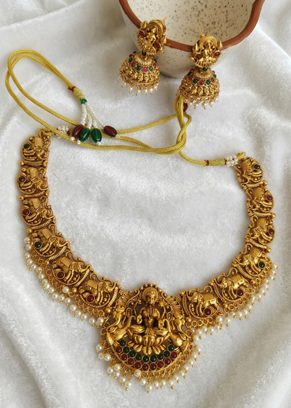An ornate gold-toned traditional necklace set with matching earrings, featuring detailed filigree, a central deity motif, and small pearl-like beads, arranged on a white textured fabric.