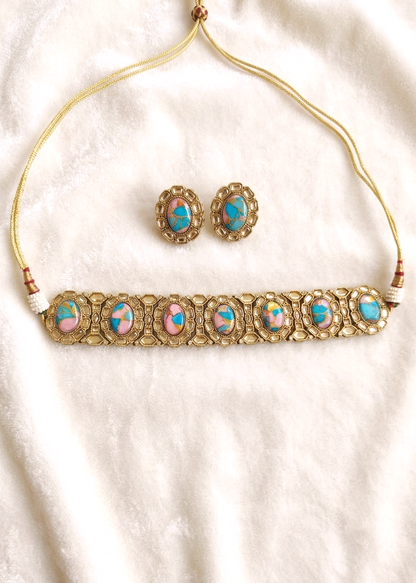 A gold-toned necklace and matching stud earrings laid flat on a soft white fabric background. The necklace has a thin gold cord and a wide ornate bar with multiple oval pink and turquoise-blue stone inlays set in gold filigree.