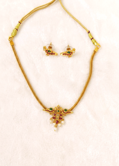 Gold-toned necklace and matching stud earrings set with small green and red stones and white pearl-like accents, displayed on a light pink textured background.