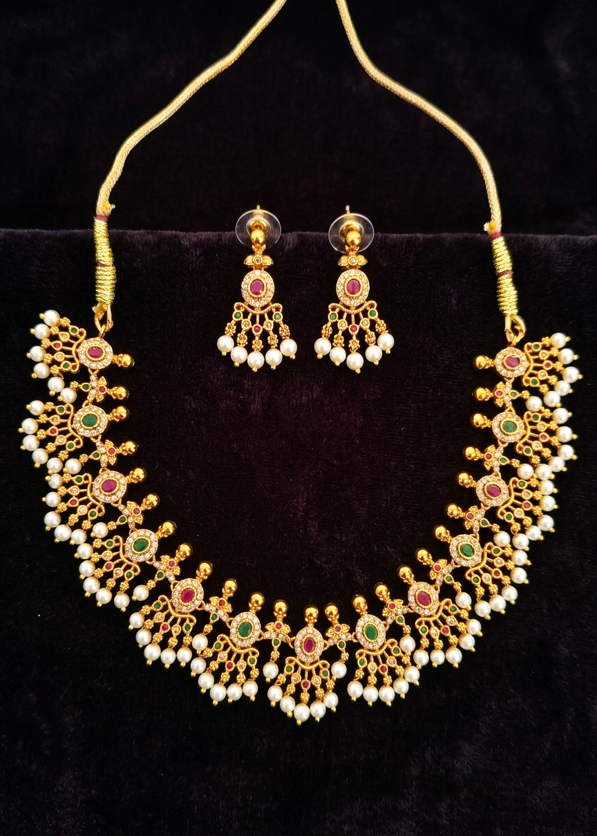 Gold-toned traditional necklace and matching drop earrings with white pearl-like beads and red and green stone accents, displayed on a black velvet surface.