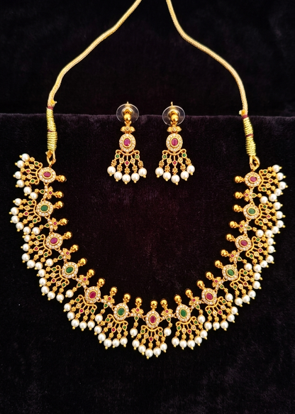 Gold-toned traditional necklace and matching drop earrings with white pearl-like beads and red and green stone accents, displayed on a black velvet surface.