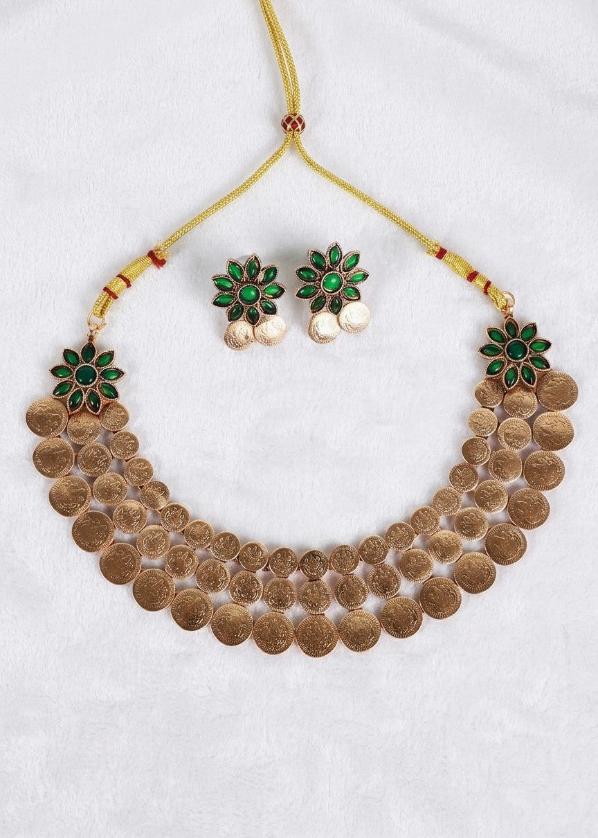 A traditional Indian gold-toned coin necklace set with matching stud earrings, featuring green and red enamel flower details and dangling white pearl-like beads, displayed on a white textured fabric background.